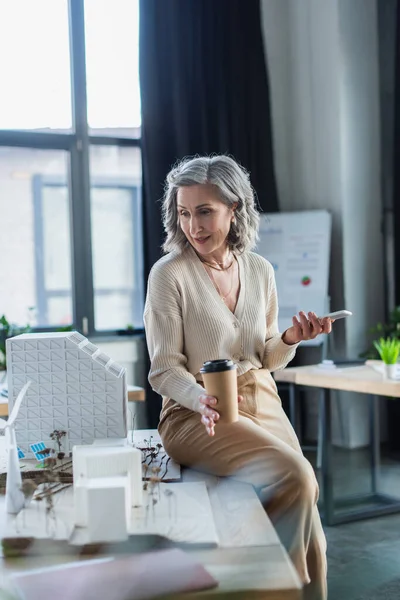 Donna d'affari in possesso di caffè per andare e telefono cellulare vicino al progetto con modelli di edifici in ufficio — Foto stock