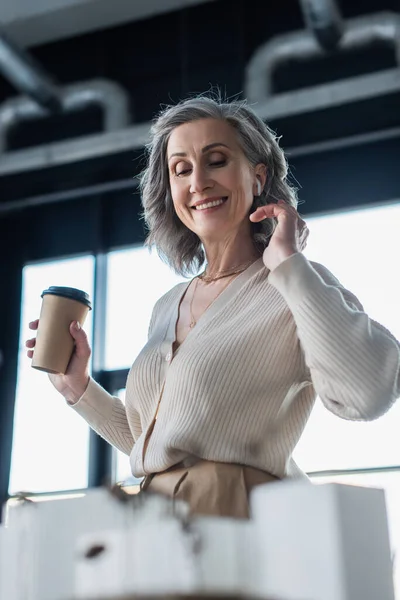 Low angle view of smiling grey haired businesswoman in earphone holding coffee to go near models of buildings in office — Stock Photo