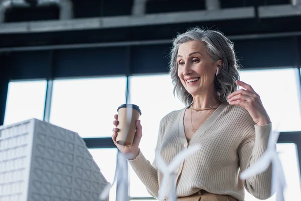 Heureuse femme d'affaires dans l'écouteur tenant boisson à emporter près du modèle flou du bâtiment dans le bureau — Photo de stock