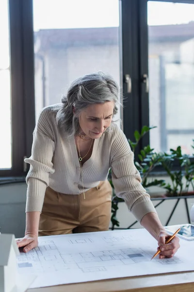 Mature businesswoman drawing on blueprint near digital tablet in office — Stock Photo