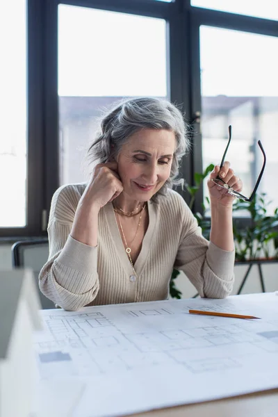 Smiling businesswoman holding eyeglasses near blueprint and model of house in office — Stock Photo