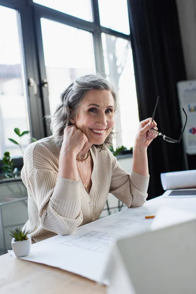 Happy businesswoman holding eyeglasses and looking at camera near blueprint in office — Stock Photo