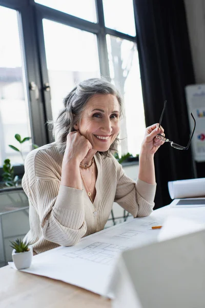 Heureuse femme d'affaires tenant des lunettes et regardant la caméra près du plan directeur dans le bureau — Photo de stock