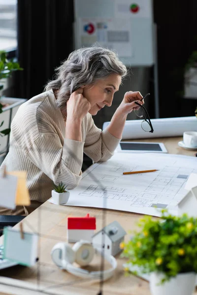 Side view of mature businesswoman working with blueprint near models of houses in office — Stock Photo