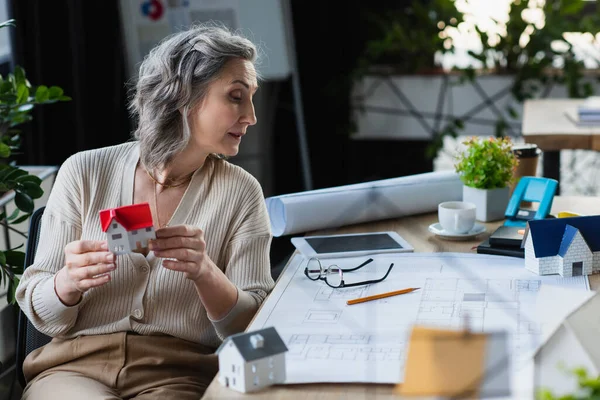 Donna d'affari matura che tiene il modello di casa vicino a cianografia e targa digitale in ufficio — Foto stock