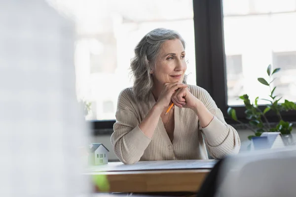 Madura mujer de negocios sosteniendo lápiz y sonriendo cerca de plano y modelos de casas en la oficina - foto de stock