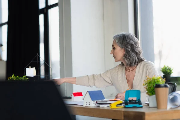 Side view of businesswoman working near models of houses and blueprints in office — Stock Photo