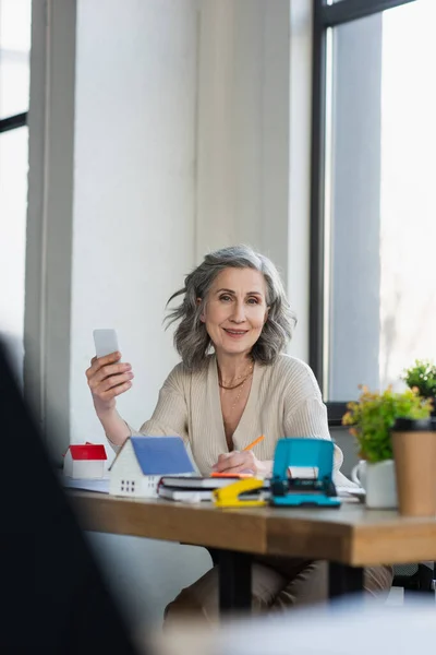 Empresária sorrindo para a câmera enquanto segura smartphone perto de modelos de casas no escritório — Fotografia de Stock