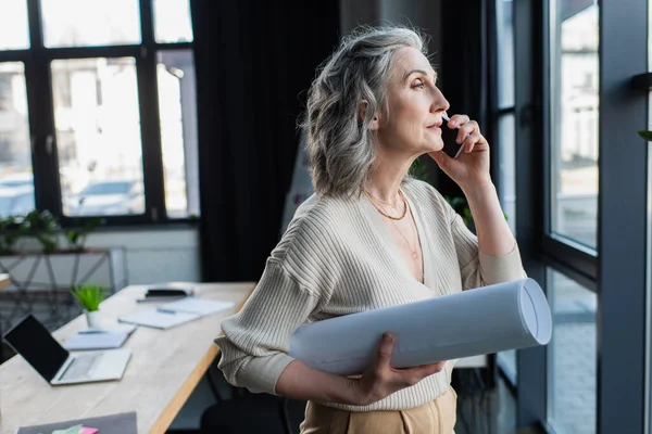 Mature businesswoman with blueprint talking on smartphone in office — Stock Photo