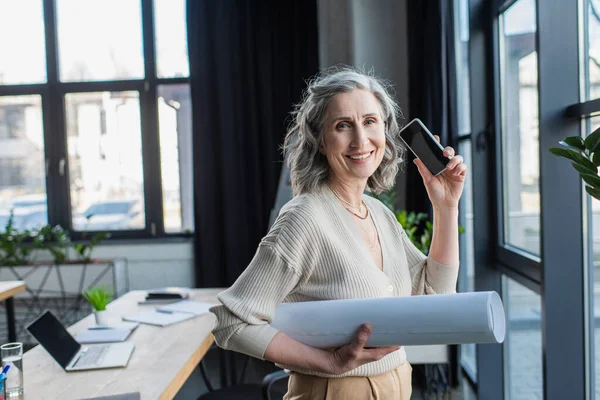 Cheerful mature businesswoman holding smartphone with blank screen and blueprint in office — Stock Photo