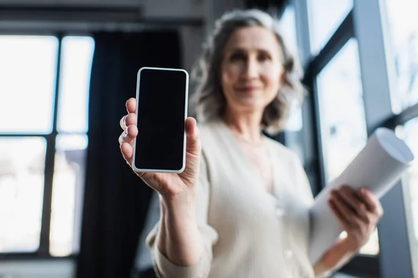 Smartphone with blank screen in hand of blurred businesswoman with blueprint in office — Stock Photo
