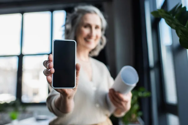 Cellphone with blank screen in hand of blurred businesswoman with blueprint in office — Stock Photo