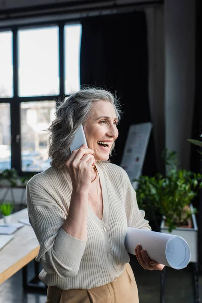 Cheerful businesswoman talking on smartphone and holding blueprint in office — Stock Photo