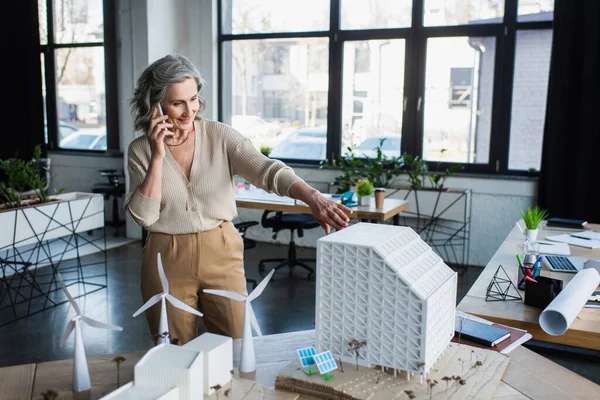 Empresária positiva falando em smartphones perto de modelos de edifícios no escritório — Fotografia de Stock