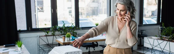 Lächelnde Geschäftsfrau spricht auf Smartphone in der Nähe Modell des Gebäudes im Büro, Banner — Stockfoto