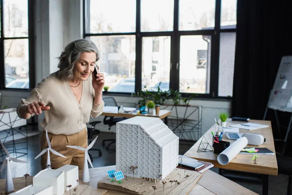 Geschäftsfrau spricht mit Smartphone in der Nähe von Gebäudemodellen und Bauplänen im Büro — Stockfoto