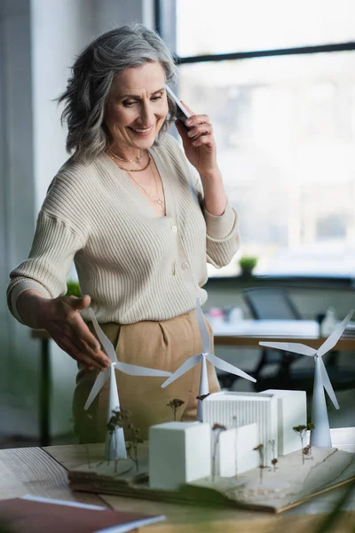 Зріла бізнес-леді розмовляє на смартфоні біля моделей будівель і паперової папки в офісі — стокове фото