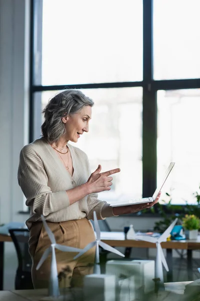 Side view of businesswoman having video call on laptop near models of buildings in office — Stock Photo