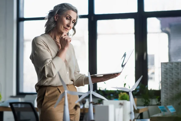 Reife Geschäftsfrau hält Laptop in der Hand und betrachtet verschwommene Modelle von Bürogebäuden — Stockfoto