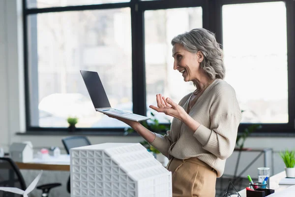 Вид сбоку позитивной бизнесвумен, имеющей видеозвонок на ноутбуке рядом с моделью здания в офисе — стоковое фото