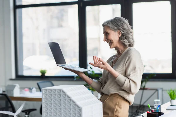 Side view of positive businesswoman having video call on laptop near model of building in office — Stock Photo