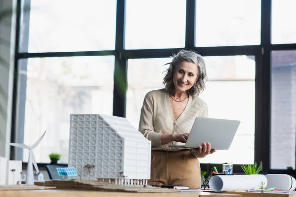 Madura mujer de negocios utilizando el ordenador portátil y mirando los modelos de edificios cerca de plano en la oficina - foto de stock