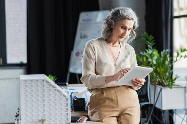 Femme d'affaires aux cheveux gris utilisant une tablette numérique près du modèle du bâtiment dans le bureau — Photo de stock