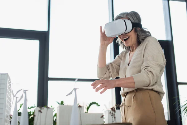 Mujer de negocios positiva en auriculares vr de pie cerca de modelos de edificios en la oficina - foto de stock