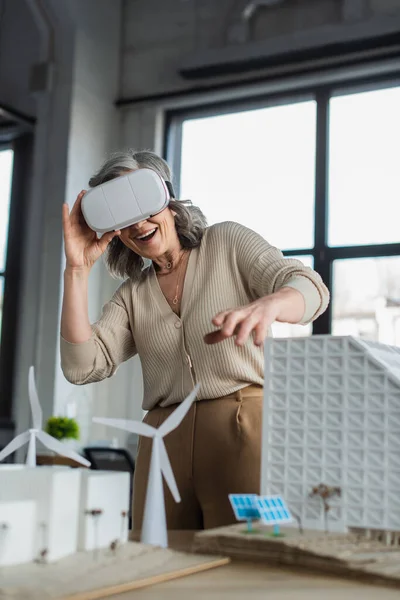 Heureuse femme d'affaires en réalité virtuelle casque près des modèles de bâtiments dans le bureau — Photo de stock