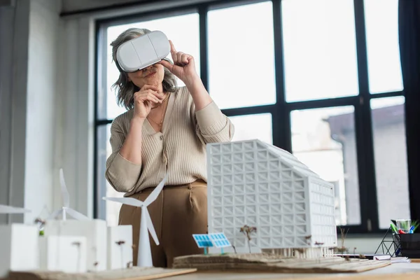 Grauhaarige Geschäftsfrau mit Kopfhörer steht in der Nähe von Modellen von Bürogebäuden — Stockfoto