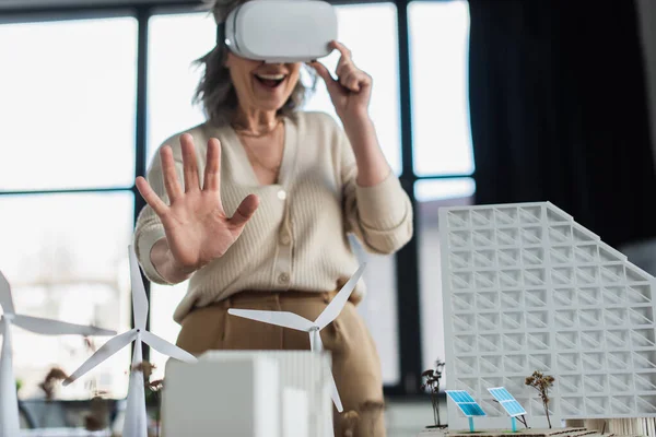 Empresaria borrosa en auriculares vr mostrando gesto de parada cerca de modelos de edificio en la oficina — Stock Photo