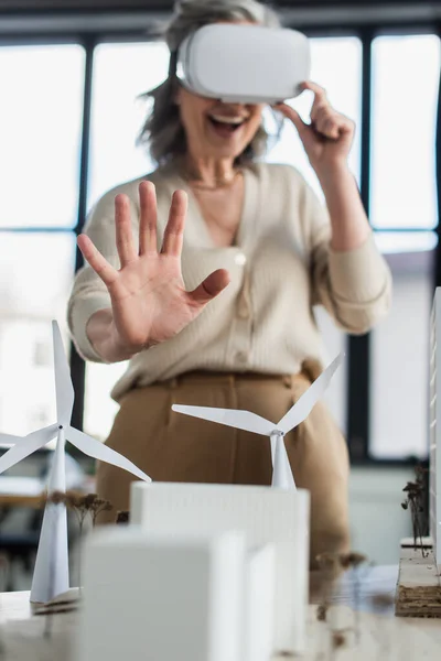 Mujer de negocios borrosa en auriculares vr que no muestran ningún gesto cerca de modelos de edificios en la oficina — Stock Photo