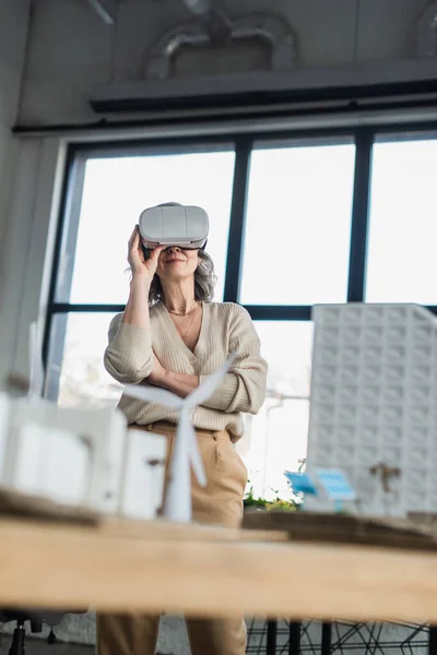 Reife Geschäftsfrau mit vr-Headset in der Nähe verschwommene Modelle von Gebäuden im Büro — Stockfoto