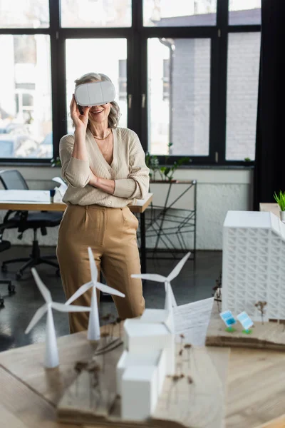 Empresária alegre em fones de ouvido de realidade virtual em pé perto do projeto com modelos de edifícios no escritório — Fotografia de Stock