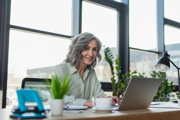 Lächelnde Geschäftsfrau mit Laptop in der Nähe von Kaffee und Schreibwaren im Büro — Stockfoto