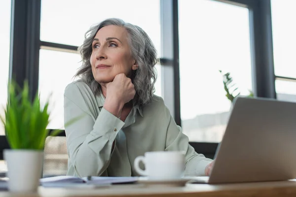 Donna d'affari matura seduta vicino a laptop sfocato e caffè in ufficio — Foto stock