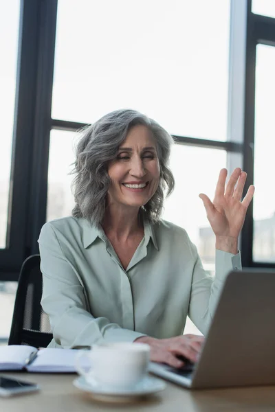 Felice donna d'affari matura avendo video chat sul computer portatile vicino notebook e caffè in ufficio — Foto stock