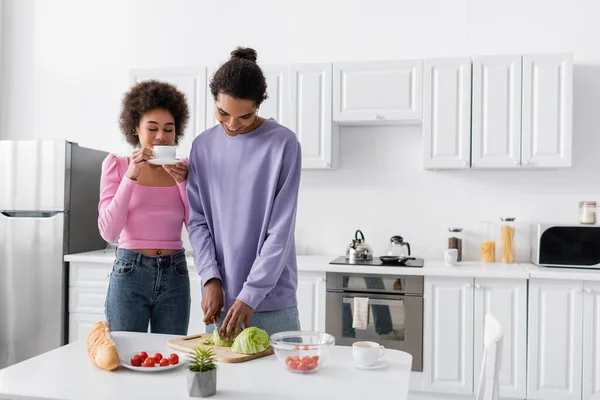 Smiling african american man cutting cabbage near girlfriend with coffee cup in kitchen — Foto stock