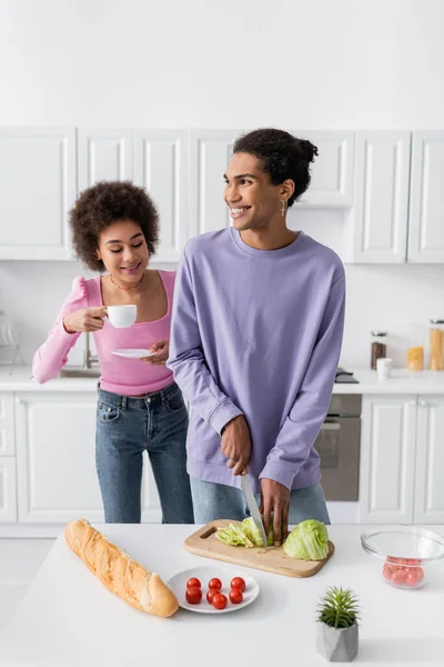 Cheerful african american woman holding cup near boyfriend cutting salad in kitchen — Stockfoto