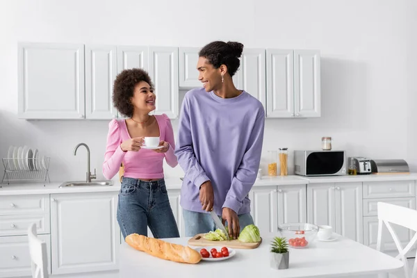 Happy african american woman with cup looking at boyfriend cooking salad in kitchen — Stockfoto