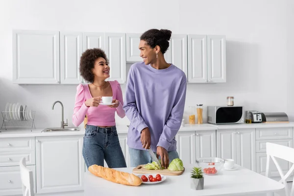 Feliz africana americana mujer con taza mirando novio cocina ensalada en cocina - foto de stock