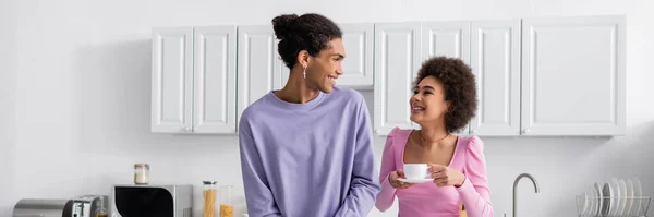 African american man looking at positive girlfriend with cup of coffee in kitchen, banner — Foto stock