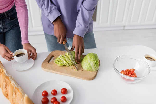 Cropped view of african american man cutting cabbage near girlfriend with coffee and baguette in kitchen — Stock Photo