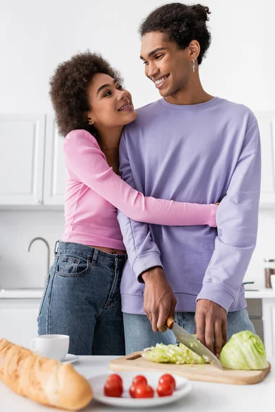 Sorridente donna africana americana abbracciare fidanzato taglio cavolo in cucina — Foto stock