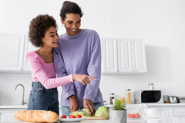 Positive Afroamerikanerin zeigt auf Kirschtomaten, während Freund zu Hause Salat kocht — Stockfoto