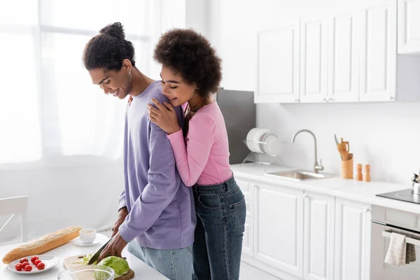 Joven mujer afroamericana abrazando novio cocina ensalada cerca de baguette y café en la cocina - foto de stock