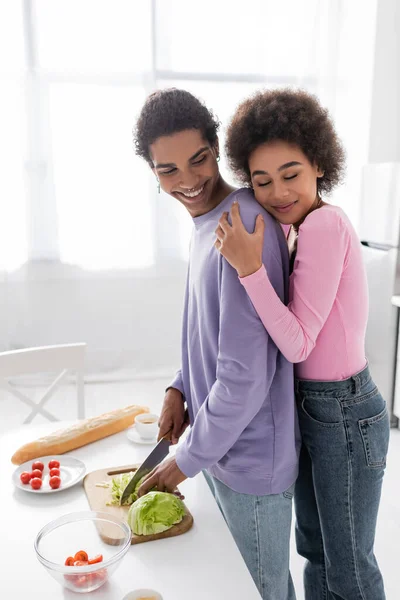 Young african american woman hugging boyfriend cooking salad at home — Foto stock