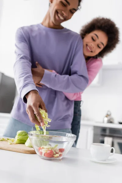 Homem Africano desfocado americano cozinhar salada perto de copo e namorada em casa — Fotografia de Stock