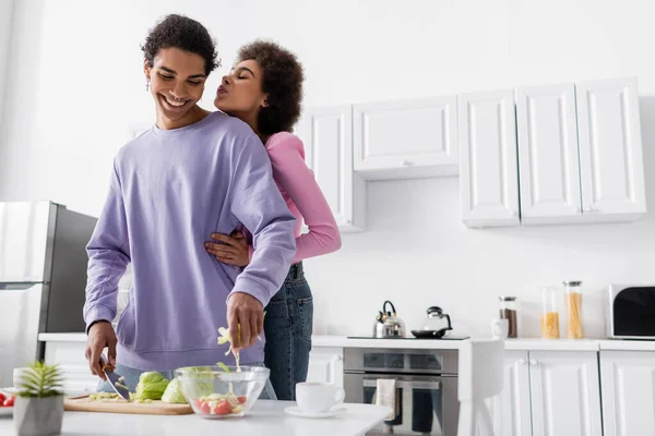 Donna afro-americana che abbraccia e brucia labbra vicino al fidanzato che cucina insalata in cucina — Foto stock