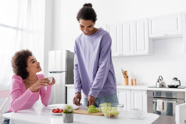 Sorridente uomo africano americano taglio insalata vicino fidanzata con una tazza di caffè a casa — Foto stock